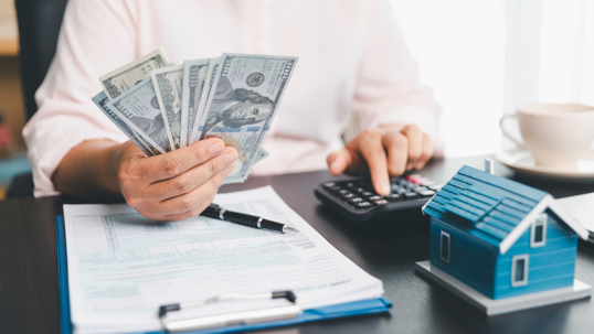 A person reviews financial documents while holding several hundred-dollar bills in one hand and using a calculator with the other. A small blue model house sits on the desk, symbolizing home expenses, mortgage planning, or real estate costs.